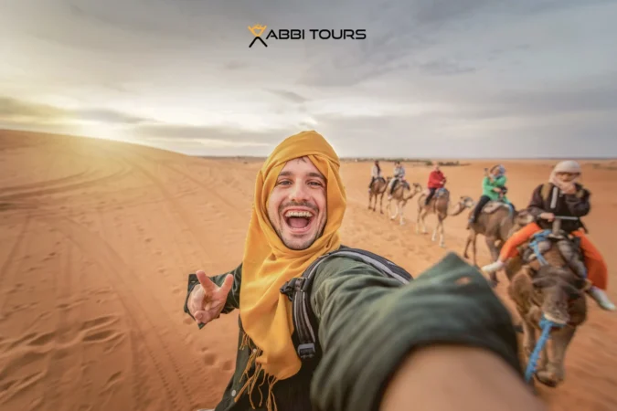 A happy man standing beside a camel in the golden sands of the Moroccan Sahara, surrounded by towering dunes under a clear blue sky.