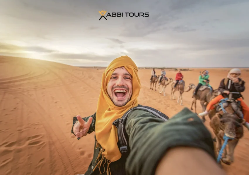 A happy man standing beside a camel in the golden sands of the Moroccan Sahara, surrounded by towering dunes under a clear blue sky.